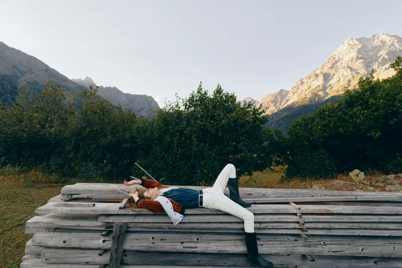 Woman reclining on stacked logs in nature with mountains in background, outdoors relax moment and landscape view, casual clothing and boots for peaceful wilderness escape.