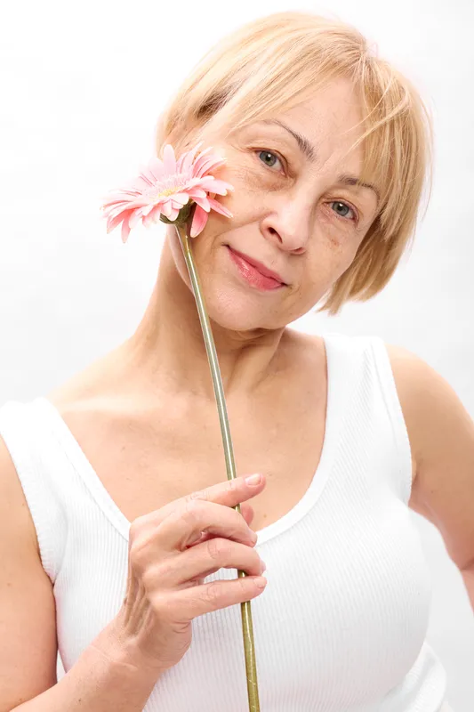 Woman, flower, elderly, smile, blonde portrait in studio on white background, pink bloom held in hands, soft lighting, gentle mood, concept of aging well and self confidence