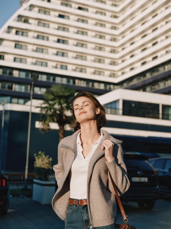 A stylish young woman stands outside a modern office building, exuding confidence and poise as she looks upward, smiles warmly, and projects a positive, aspirational vibe for passersby and colleagues.