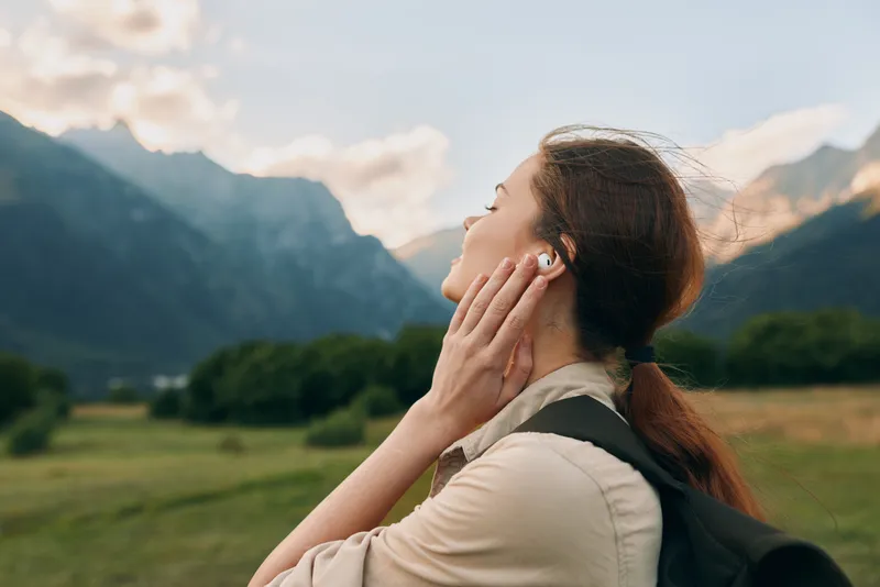 Young woman outdoors enjoying a gentle breeze in a sunlit mountain valley with distant peaks and a serene smile