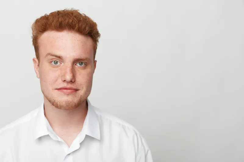 Portrait of a young redhead man with freckles wearing a white shirt looking at camera on a plain light background. Concept for casual style, confidence, and natural appearance.