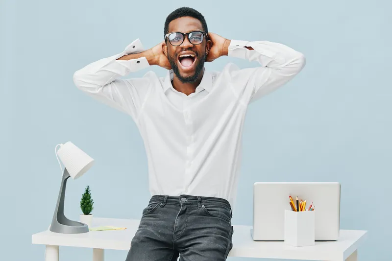 Smiling happy young man wearing glasses and white shirt, standing relaxed at modern workspace with laptop, desk lamp, plant and stationery on light blue background, studio portrait. People lifestyle c