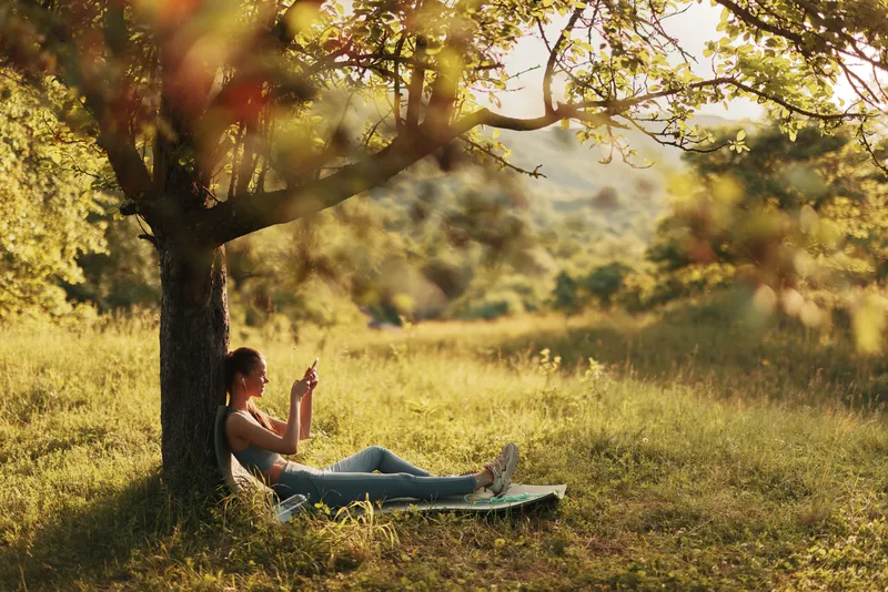 Young woman enjoying a moment of relaxation under a tree, using her smartphone in a serene outdoor setting, surrounded by nature during golden hour