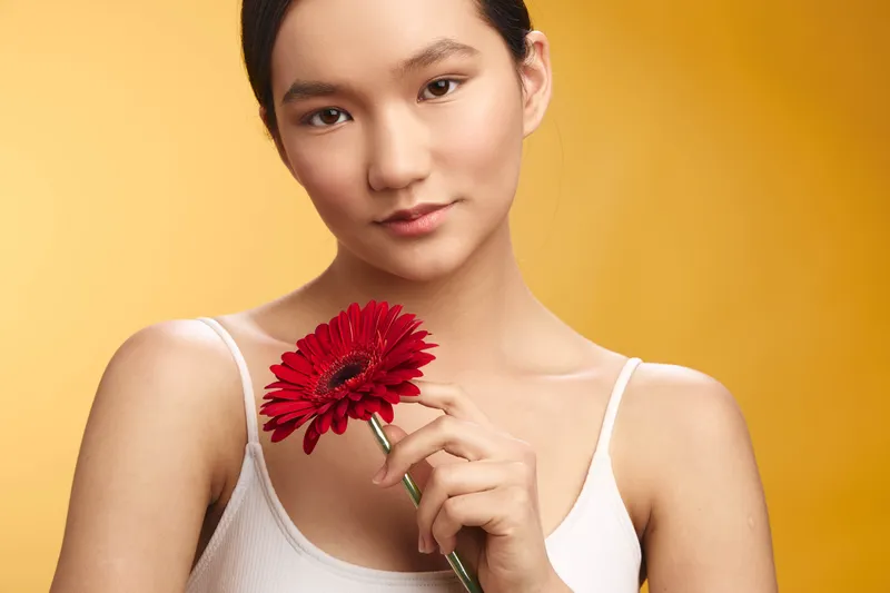 Asian woman holds a red flower against a warm yellow backdrop, presenting a serene portrait with soft lighting. The image conveys calm elegance and gentle charm.