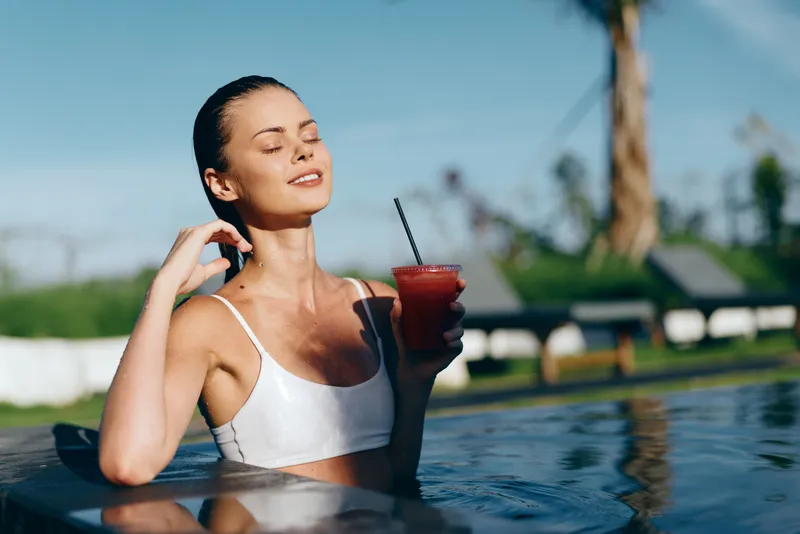A relaxed woman in a white swimsuit enjoys a red cocktail by the pool under the sun, eyes closed, embracing a calm and peaceful lifestyle moment outdoors.
