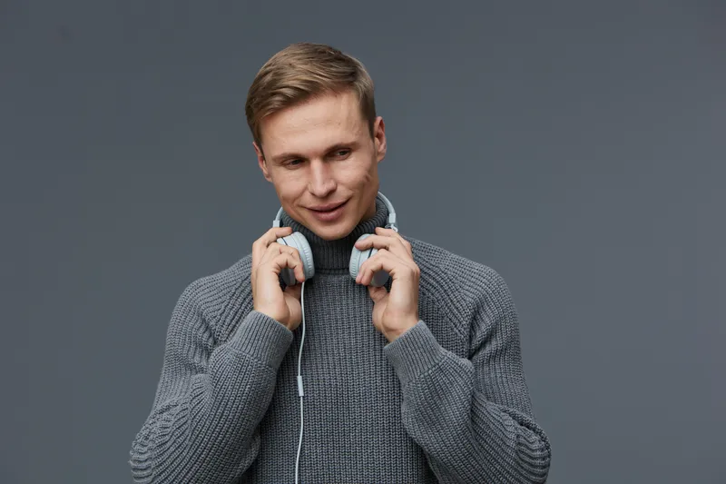 Smiling man with headphones, casual gray sweater, looking confident and relaxed, indoors against a plain gray background. Modern lifestyle, leisure, music concept, young adult male, comfortable clothi