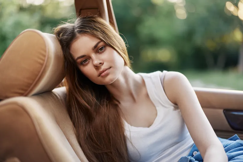 Young woman relaxing in a car during summer, with long hair and casual attire, exuding calmness in a green outdoor setting.