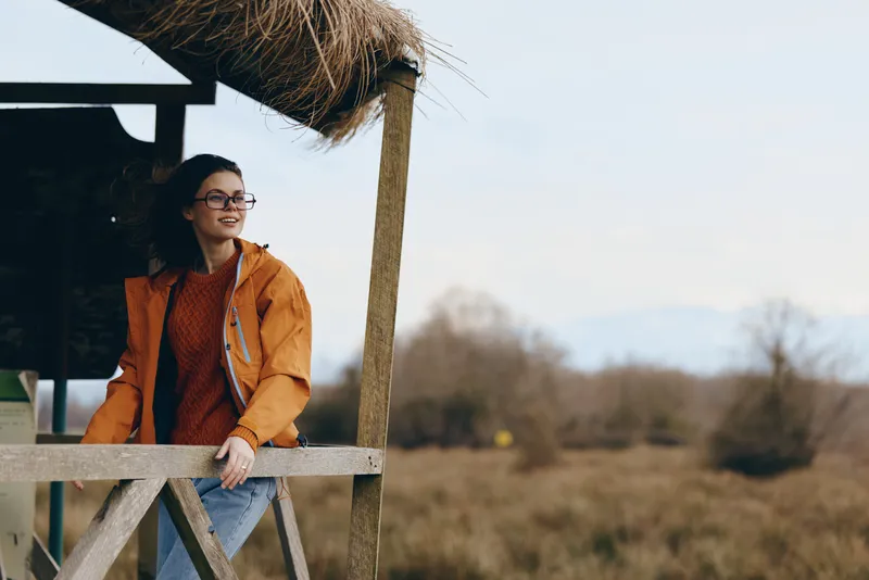 Woman lifestyle outdoors, orange jacket, sunglasses, relaxed posture on wooden deck, sunlight and soft breeze, calm smiling expression, countryside background with trees and sky blur