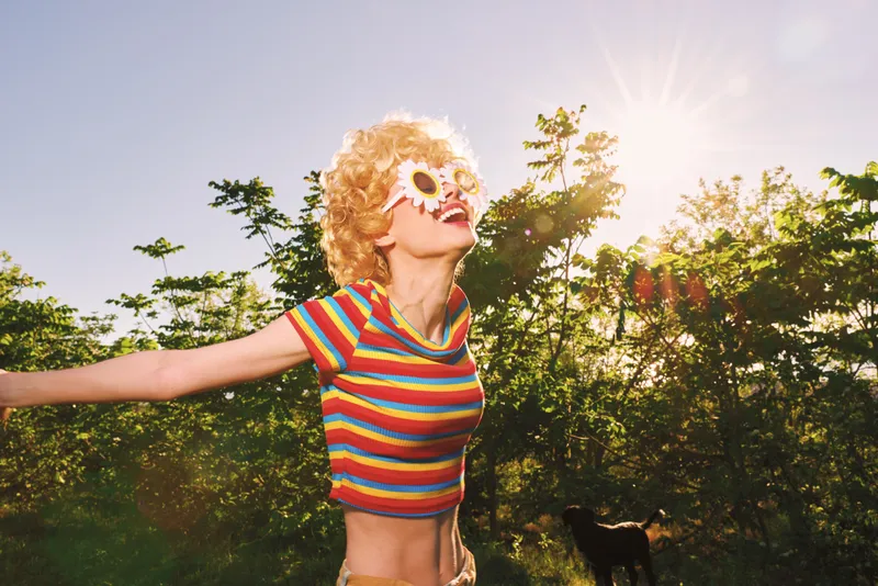 Woman enjoys a sunny outdoor moment with sunflower sunglasses, wearing a colorful striped top, arms open, radiant smile, carefree mood, bright natural light, nature backdrop