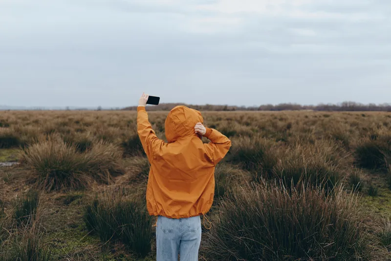 Woman outdoors in a grassy field, wearing orange jacket with hood, standing with arms raised holding a small flag, lifestyle moment, freedom and happiness, cloudy light, outdoor calm