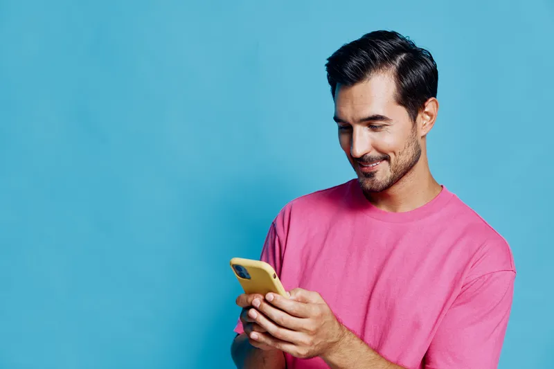 Smiling man wearing pink t-shirt looks at his mobile phone, enjoying a moment of connection, captured against a solid blue background. Modern lifestyle concept.