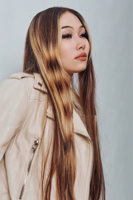 Young woman with long straight hair wearing beige leather jacket posing with calm expression against light wall indoor. Fashion portrait with natural makeup and confident look for style concept.
