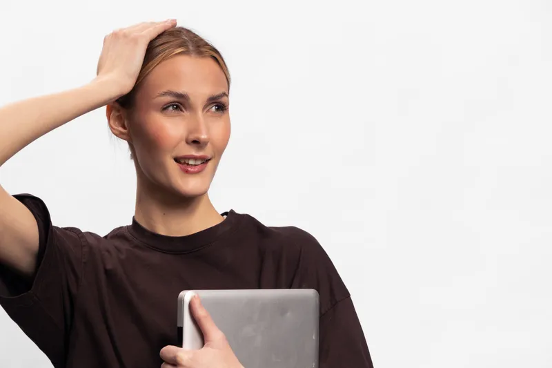 Young woman in casual brown shirt holds laptop, looks thoughtful with hand on head. Isolated on white background. Concept of technology, contemplation, and casual lifestyle.