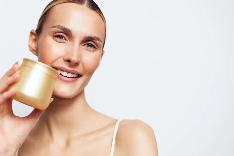 A young woman with a soft smile holds a gold cosmetic jar in front of a clean white background, presenting a skincare routine and self care concept.