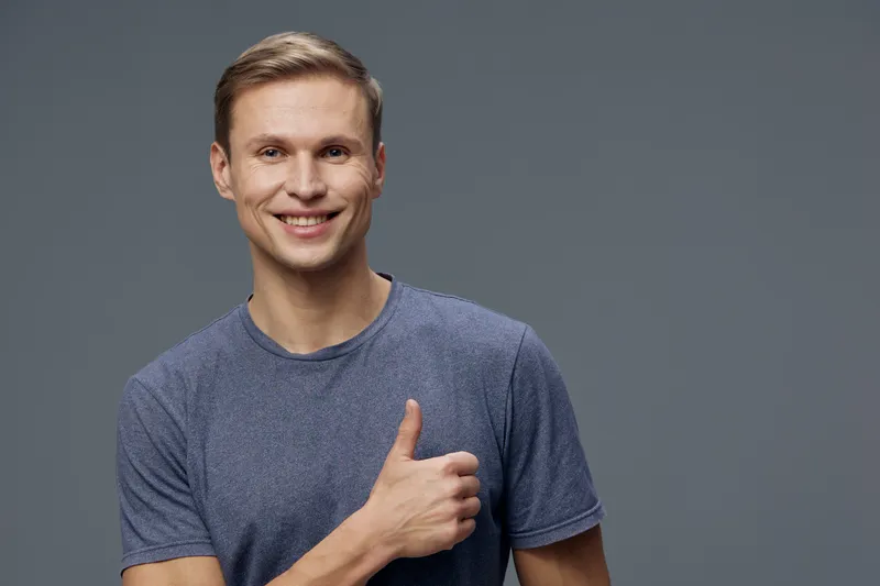 Smiling man in casual blue t-shirt on plain grey background, looking at camera, making thumbs-up gesture, happy, relaxed, friendly, positive vibe, studio portrait.