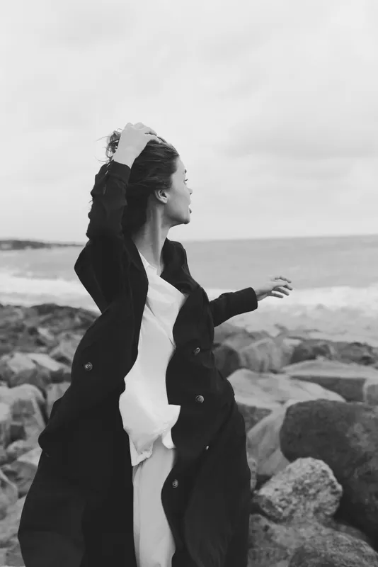Wind swept figure on a rocky shore in a monochrome coat, catching sea breeze with dynamic movement, dramatic mood, and timeless coastal atmosphere