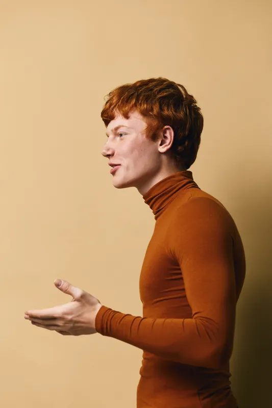 Young man with red hair wearing brown turtleneck standing sideways and speaking with hand gestures, isolated on beige background with casual expression and profile view.