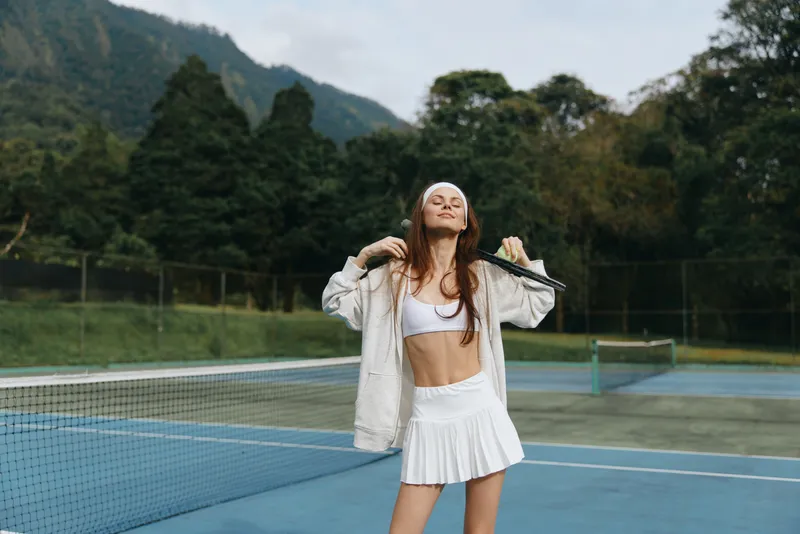 Young female tennis player in white sportswear smiling on the court with a racket, showcasing confidence and vitality against a natural green background