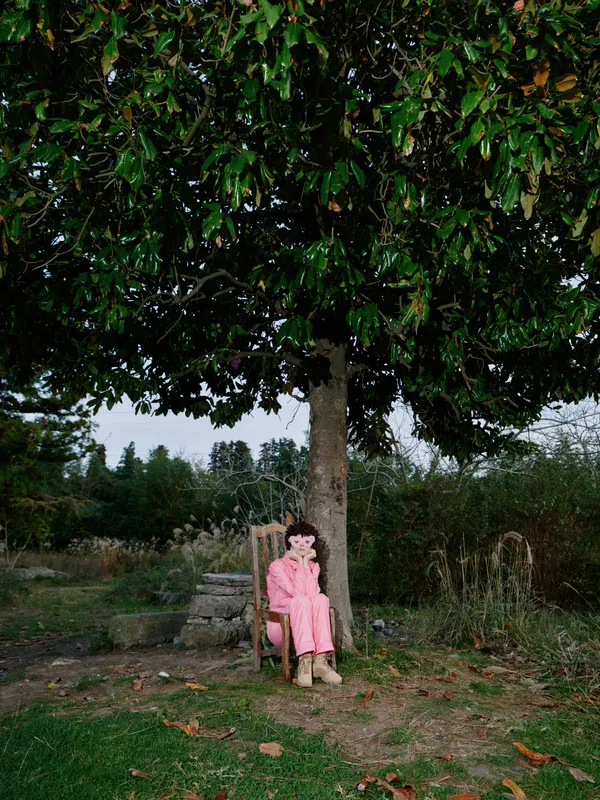 A young girl sits on a wooden bench beneath a large leafy tree in a quiet park, wearing a pink coat and boots, surrounded by grass, fallen leaves and expansive calm space.