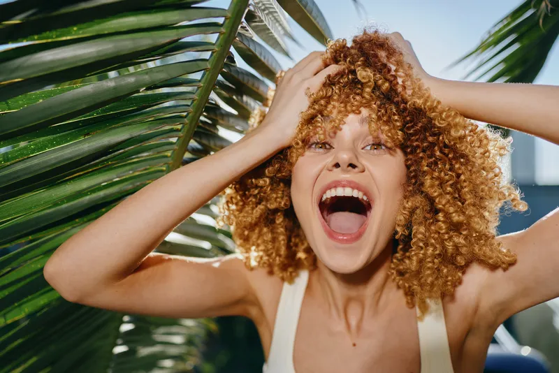 excited woman with curly hair joyfully laughing outdoors near large tropical leaves in bright sunlight, summer vibe and natural energy