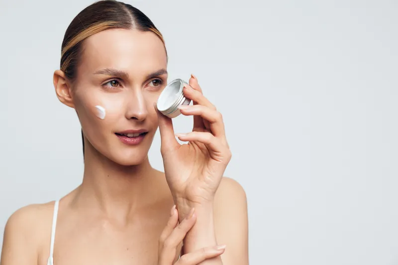 Young woman with blonde hair applies face cream on her cheek while holding an open jar in a bright studio, showing skincare routine and natural beauty care.