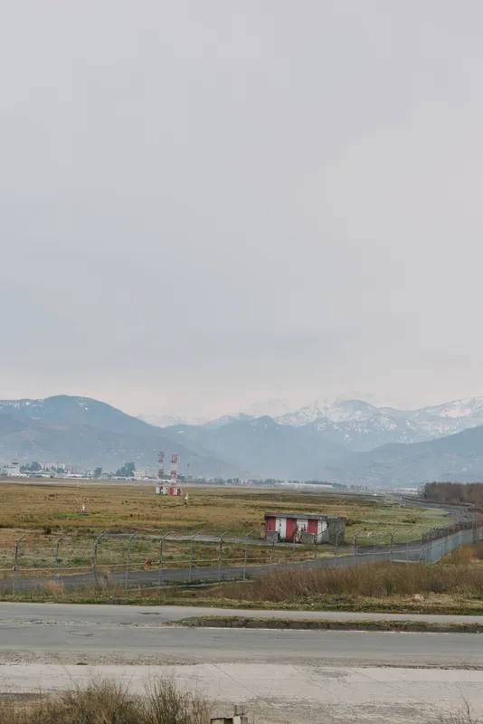 Landscape with grassy field and small buildings under a cloudy sky. Mountains are visible in the distance, creating a calm and natural outdoor environment scene.