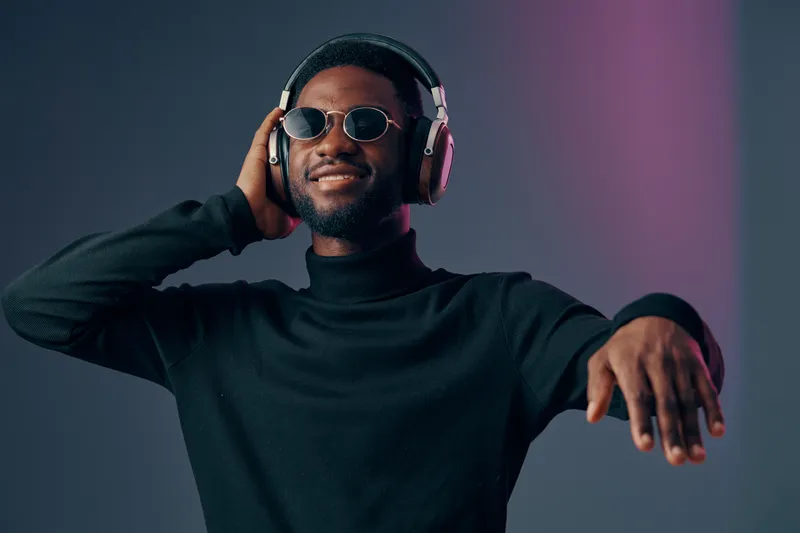 Young man in black turtleneck and sunglasses enjoying music with headphones, showcasing a joyful expression in a modern studio backdrop with soft purple lighting