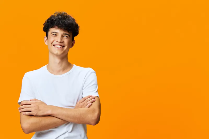 smiling man with curly hair wearing white t-shirt and crossed arms stands confidently against vibrant orange background, radiating positivity and youthfulness