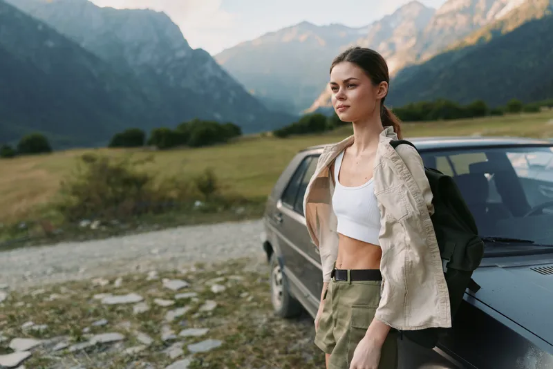 Young woman stands beside a car in a mountain valley, looking ahead with calm confidence and relaxed posture in a sunlit, open landscape with distant peaks.