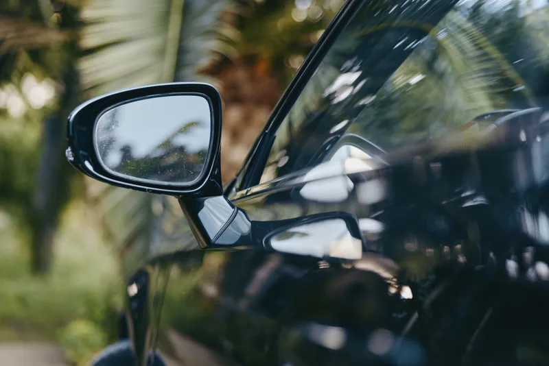 Car side mirror of black car with rain droplets, tropical background and glossy reflection; closeup detail of sleek luxury sedan exterior for travel and lifestyle imagery.