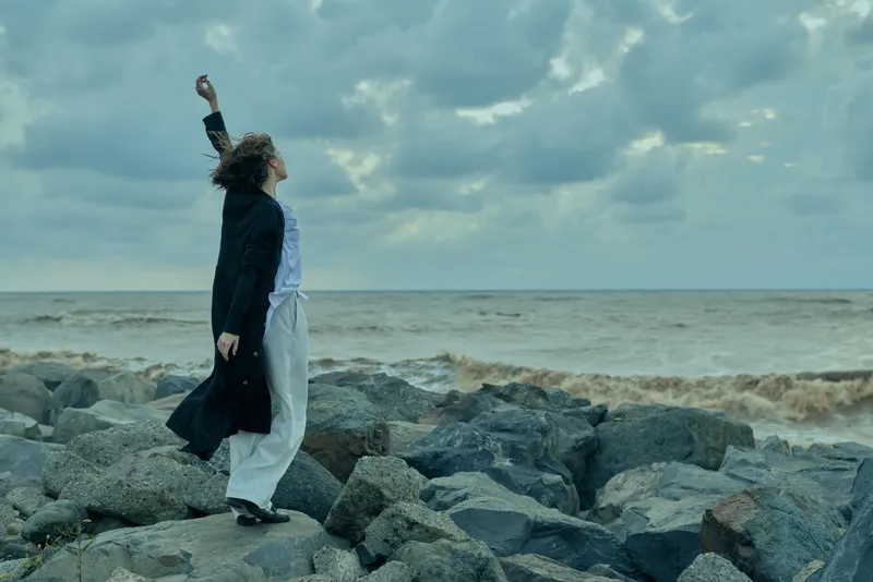 Sea, rocks, wind, couple, outdoors, shoreline, cloudy, movement describe two figures on rugged coast, standing with raised posture as coats billow under a stormy horizon