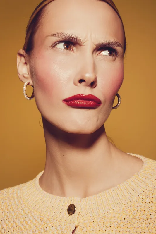 Close-up of a woman with red lipstick and gold hoop earrings making a skeptical facial expression against a warm yellow background. Concept of doubt and attitude.