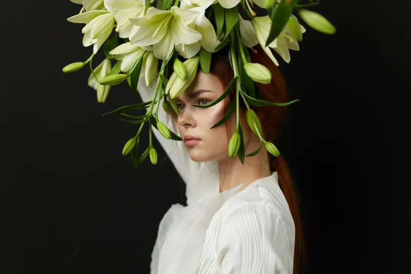 Elegant young woman with long red hair wearing a white dress and a large floral arrangement of white lilies on a dark background, embodying beauty and nature