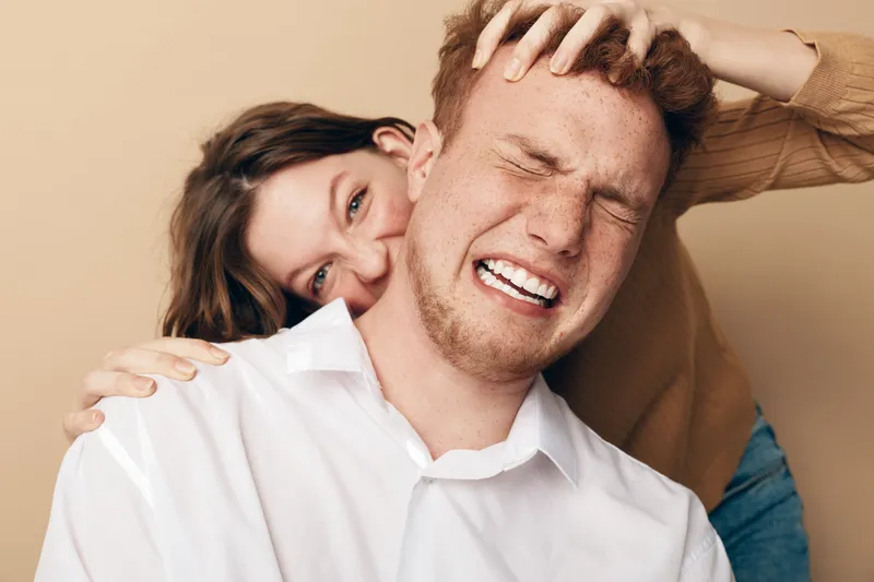 Playful young woman with brown hair teasing redhead man in white shirt, who grimaces with eyes closed. Casual indoor portrait on beige background, concept of fun, friendship, and humor.