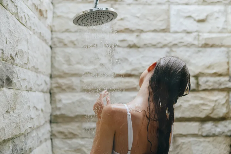 Woman taking outdoor shower with wet hair and stone wall background, enjoying refreshing water under natural light in warm weather and casual white swimwear.