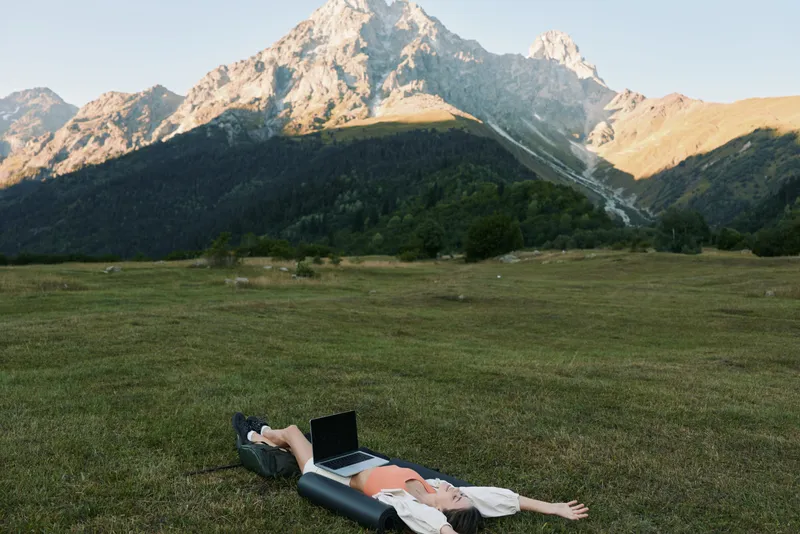 A person lies on a grassy meadow with a laptop, surrounded by towering mountain peaks. The scene conveys outdoor work, calm focus, and tranquil productivity in nature.