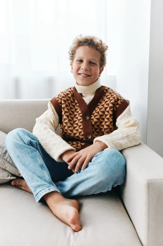 Smiling boy sits relaxed on a light sofa wearing a warm patterned knitted vest over a turtleneck and jeans, capturing a cozy home portrait.