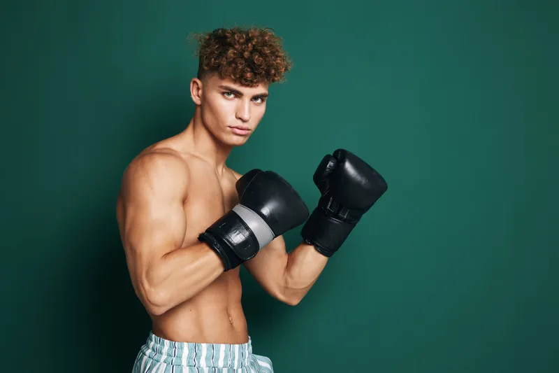 Young male boxer with curly hair wearing black gloves and striped shorts posing in fighting stance against green background for fitness and sports training.