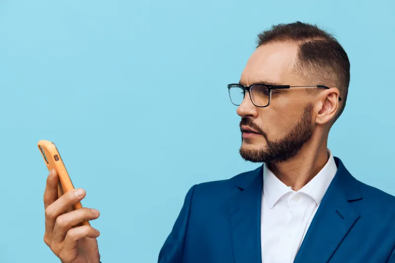 A professional business man in a sharp blue suit checks his smartphone, displaying focused and confident emotions against a solid color background.