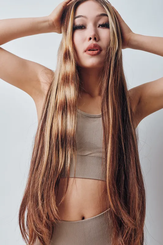 Young woman with long straight hair posing in beige crop top and pants, looking at camera with hands in hair against plain background, beauty and fashion concept.
