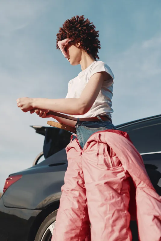 Casual woman outdoors beside a car, wearing a white tee and pink pants, captured in bright daylight with a clear sky, conveying a relaxed road trip mood and everyday style