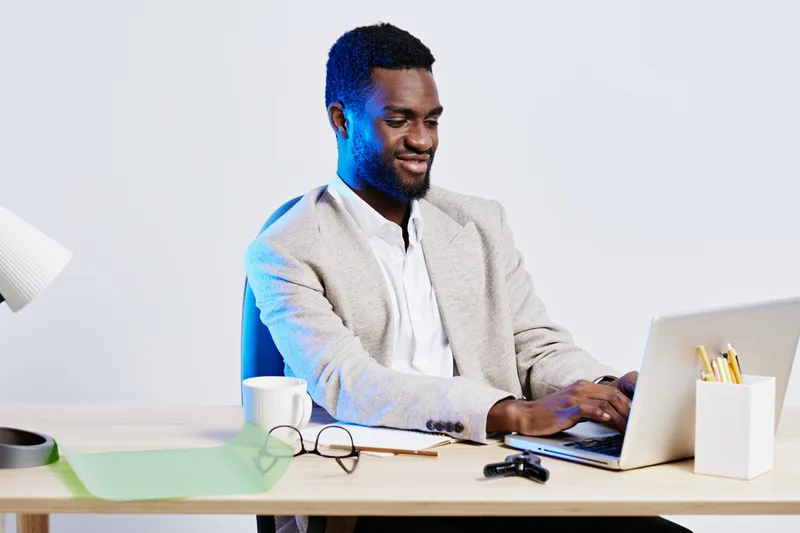 Man working on laptop at desk in modern office, smiling, using computer, indoors, professional business environment, casual attire, focused, mid adult male, workplace productivity, concept: remote wor