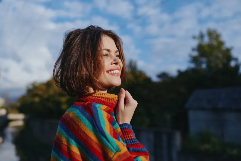 Smiling woman portrait in colorful sweater outdoors, side profile with short hair and sunlight, happy expression enjoying nature and breeze, casual fashion and warm mood.