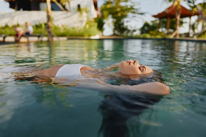 Relaxed woman floating on water surface in outdoor pool, eyes closed, peaceful expression, sunny day, calm atmosphere, natural environment, lifestyle concept