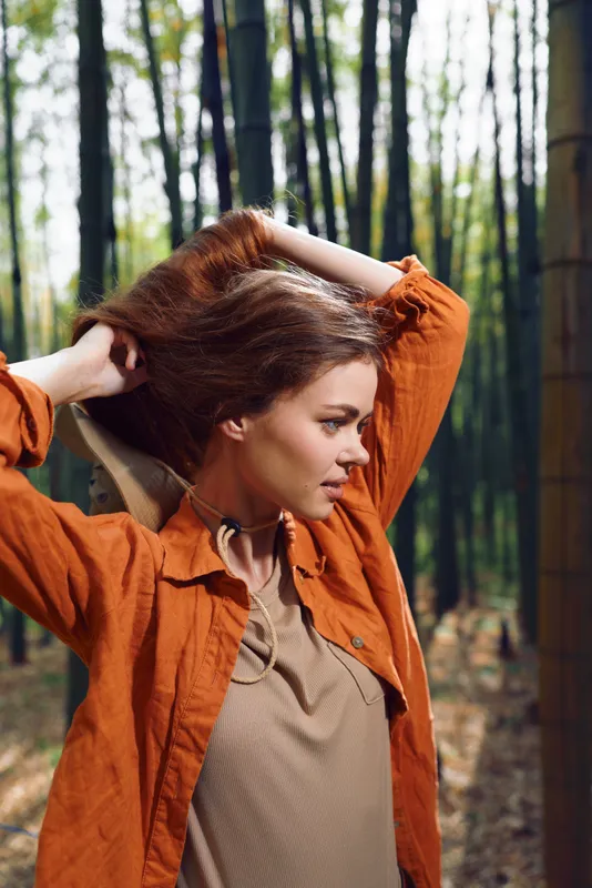 Woman portrait in forest, profile view with long hair and rust jacket, outdoors lifestyle photo of young female model posing among tall bamboo trees with natural sunlight and serene mood