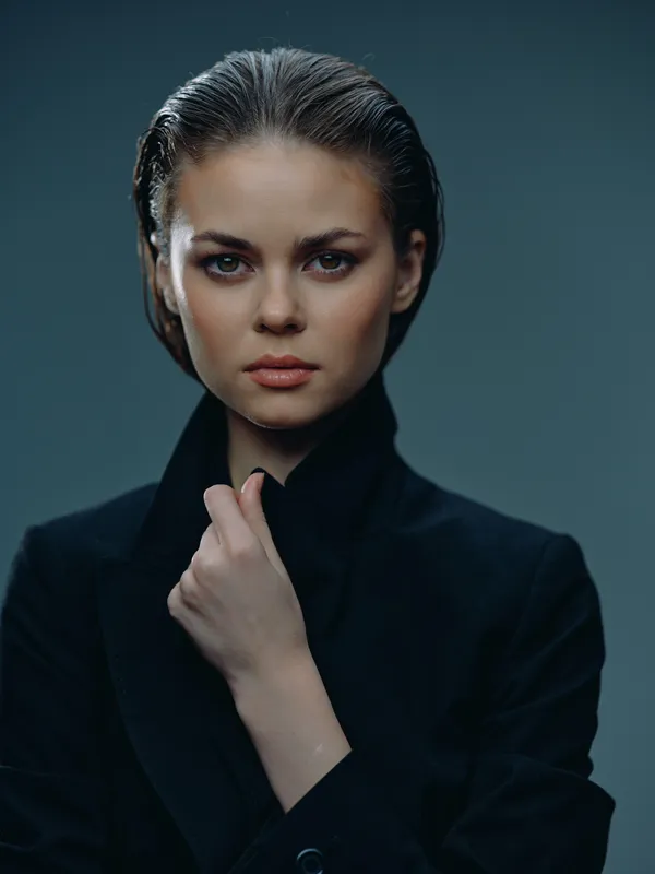 serious woman with slicked-back hair wearing a black blazer, confident and intense expression in a dark studio setting