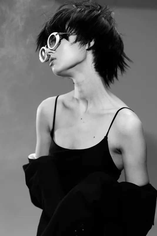 Black and white portrait of a stylish woman wearing round glasses and a black outfit with short hair and a confident pose.