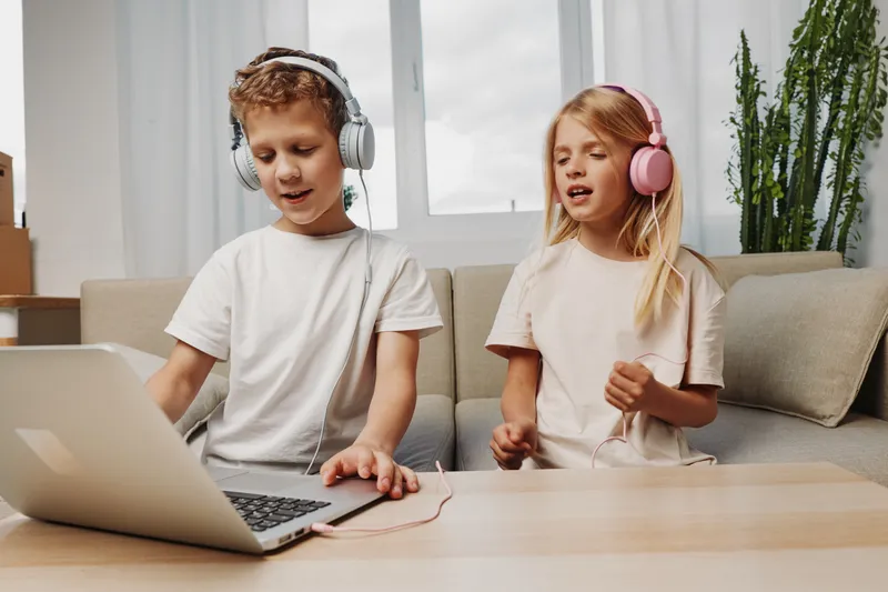 Two children wearing headphones enjoy singing and playing music together using laptop in cozy living room with natural light and casual outfits. Concept of creativity and childhood fun.