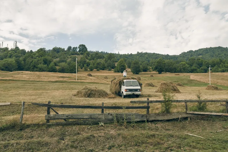 Rural countryside scene featuring a white van in a field of hay, with a wooden fence in the foreground, rolling hills and a cloudy sky.