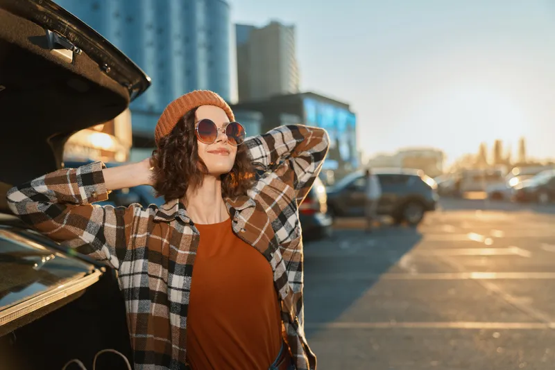 Woman by car in parking lot with sunlight, sunglasses and beanie, relaxed candid pose capturing authenticity, mindful living and emotional storytelling in a warm golden hour glow urban scene.